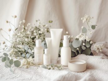 several white containers reside alongside delicate white flowers and soft eucalyptus leaves on a towel
